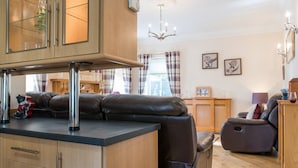 Een moderne keuken met een vast kookeiland, een eetgedeelte met een houten tafel en stoelen, en een woonkamer met een bank en een massagestoel.