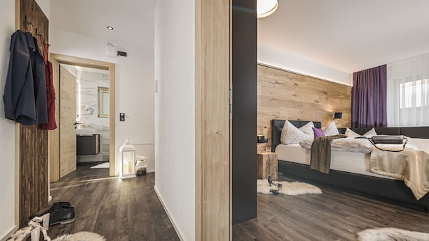 A modern bedroom with a wooden headboard, a large bed with multiple pillows, a nightstand, and a bedside table. There is a mirror on the wall and a door leading to another room. - 2 bedrooms, kitchen, bathroom, next to the valley run and the valley station (Kaltenbach)