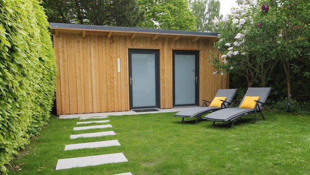 Eine Holzhütte mit zwei Türen, ein Garten mit einem Kiesweg und zwei Liegestühle mit gelben Kissen.