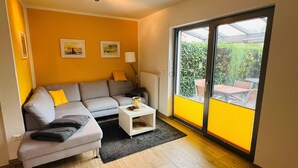 Ein modernes Wohnzimmer mit einem grauen Sofa, einem kleinen weißen Beistelltisch, einem schwarzen Teppich und einem großen Fenster mit gelben Jalousien.