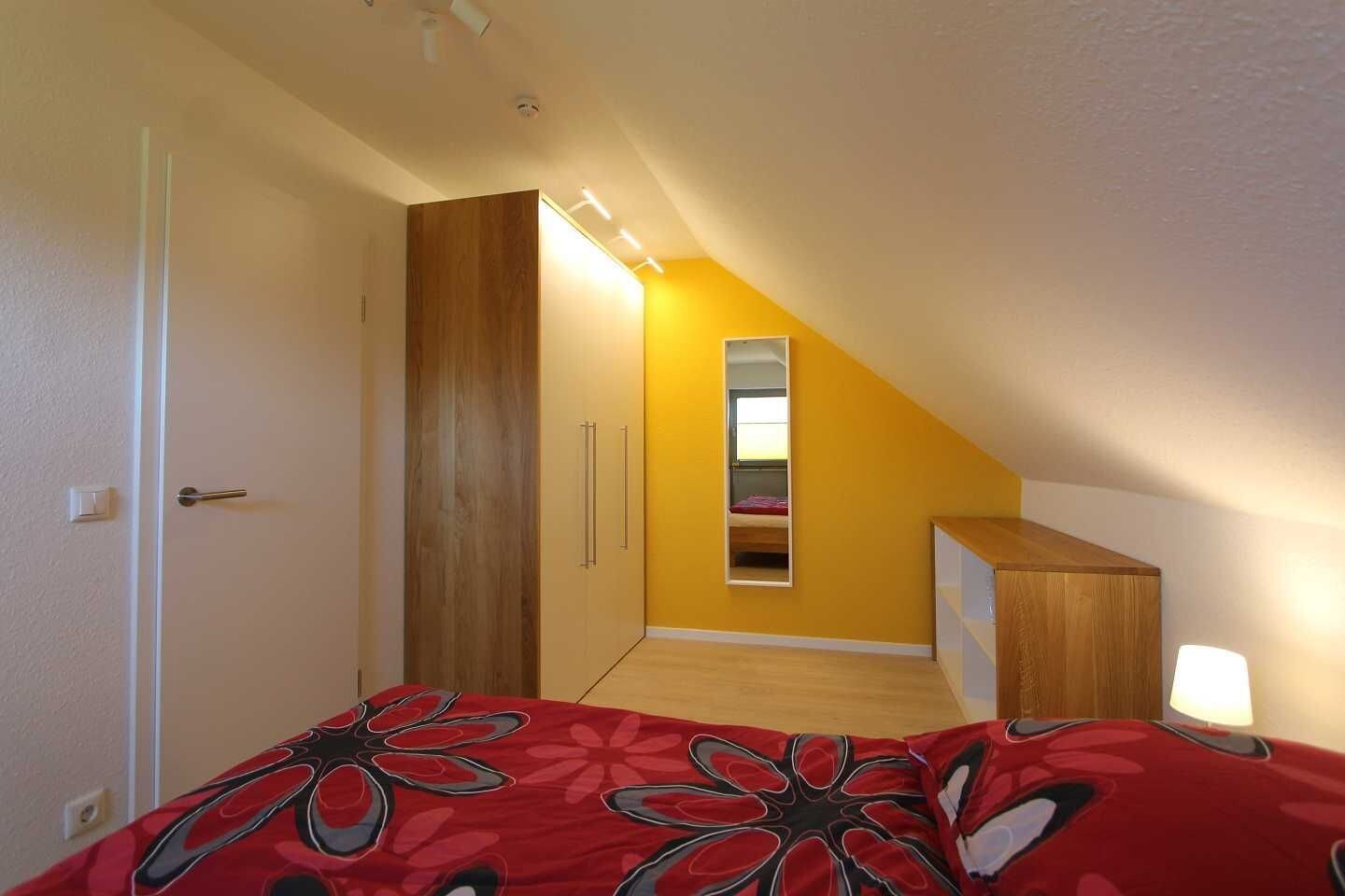 Een slaapkamer met een schuine kap, een bed met een rode bloemenovertrek, een houten garderobe, een nachtkastje met een lamp en een spiegel aan de muur.