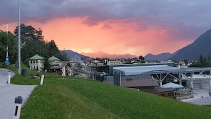 Une ville où se côtoient des bâtiments traditionnels et modernes, une rue principale animée avec un feu de signalisation, et un paysage de montagnes se découpant sur un ciel aux couleurs vives.