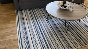 Een woonkamer met een grijze bank, een blauw-wit tapijt met dessin en een ronde salontafel met decoratieve objecten.