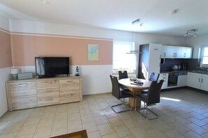 A modern kitchen with white cabinets, a dining table with chairs, a television on a wooden cabinet, and a window providing natural light. - Leuchtturmstraße house 15, only 150 m from the beach - Leuchtturmstraße 31 (Rerik)