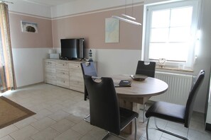 A dining area with a round table, two black chairs, a television on a cabinet, and a window with blinds. - Leuchtturmstraße house 15, only 150 m from the beach - Leuchtturmstraße 31 (Rerik)