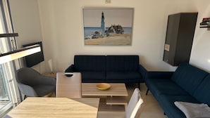 Una sala moderna con un sofá azul oscuro, una mesa de centro de madera y una mesa de comedor con sillas. Un cuadro de un faro está en la pared.