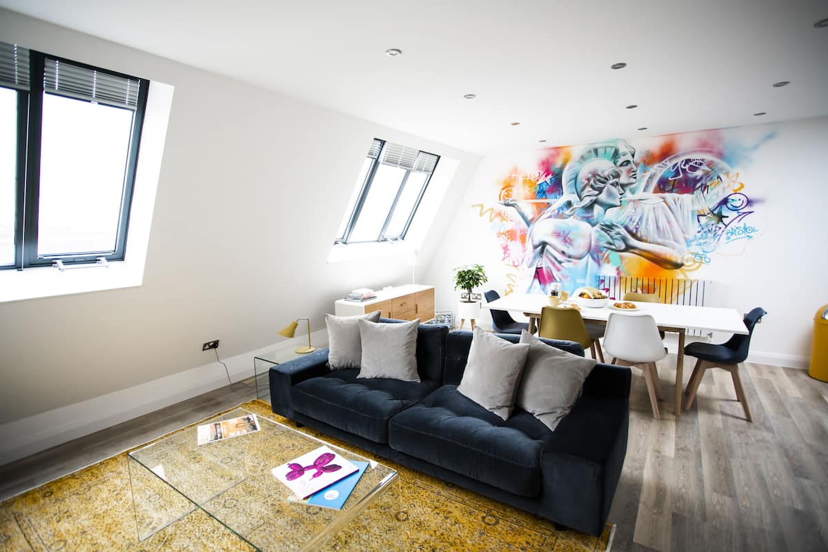 Ein modernes Wohnzimmer mit einem dunkelblauen Sofa, einem Glastisch und einem farbenfrohen Graffiti-Wandbild.