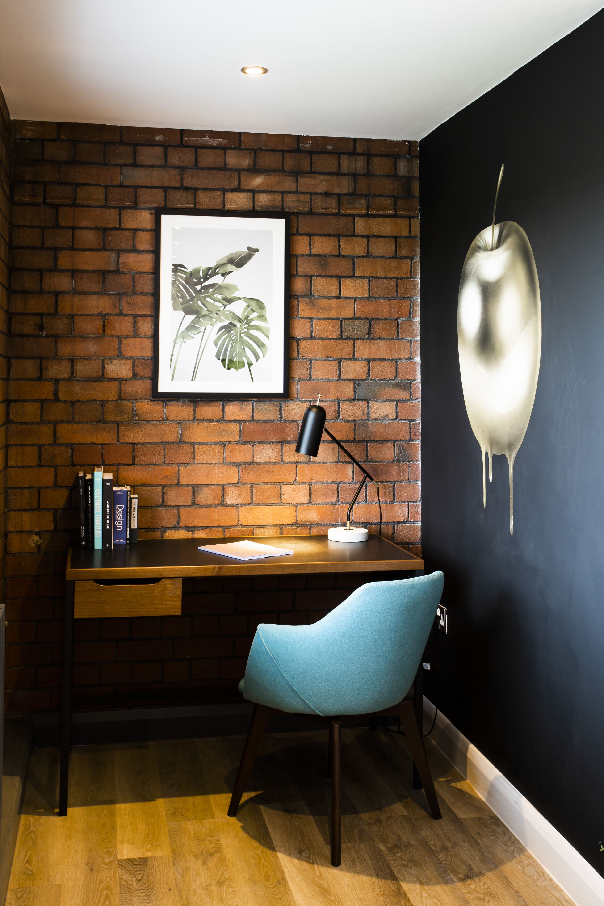 A room with a brick wall, a desk with a chair, a framed plant, and a wall painting.