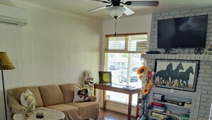 A living room with a sofa, a round wooden coffee table, a television mounted on a brick wall, and a window with blinds.
