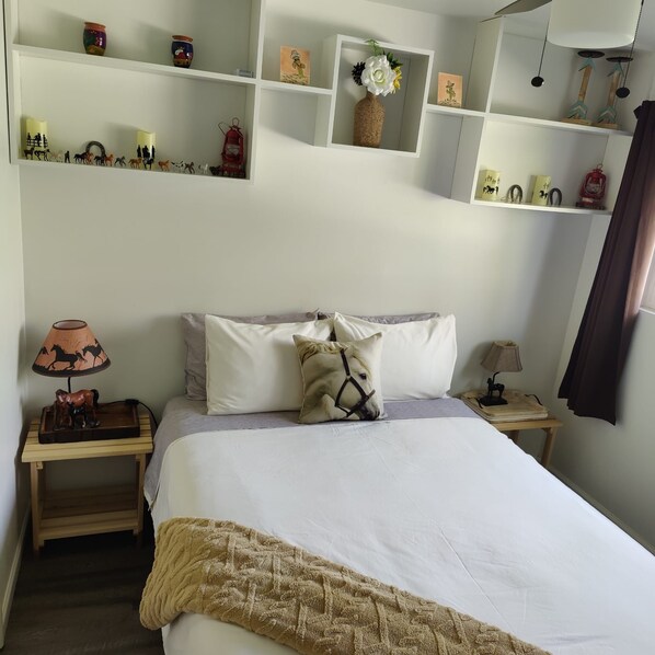 A neatly arranged bedroom with a bed, pillows, a nightstand, a lamp, and shelves with various items.