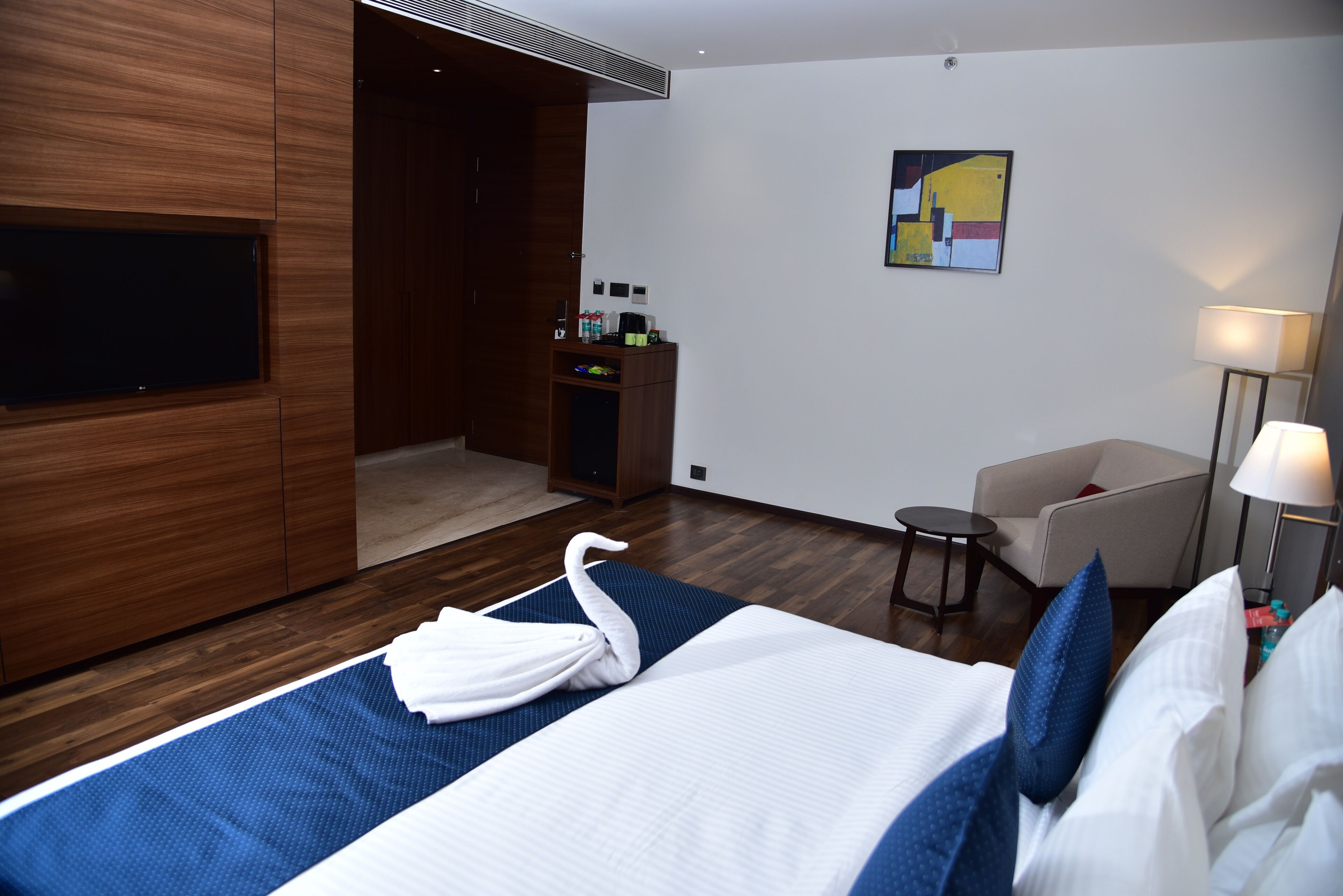 A hotel room with a bed, a TV, a desk with a lamp, and a painting on the wall.
