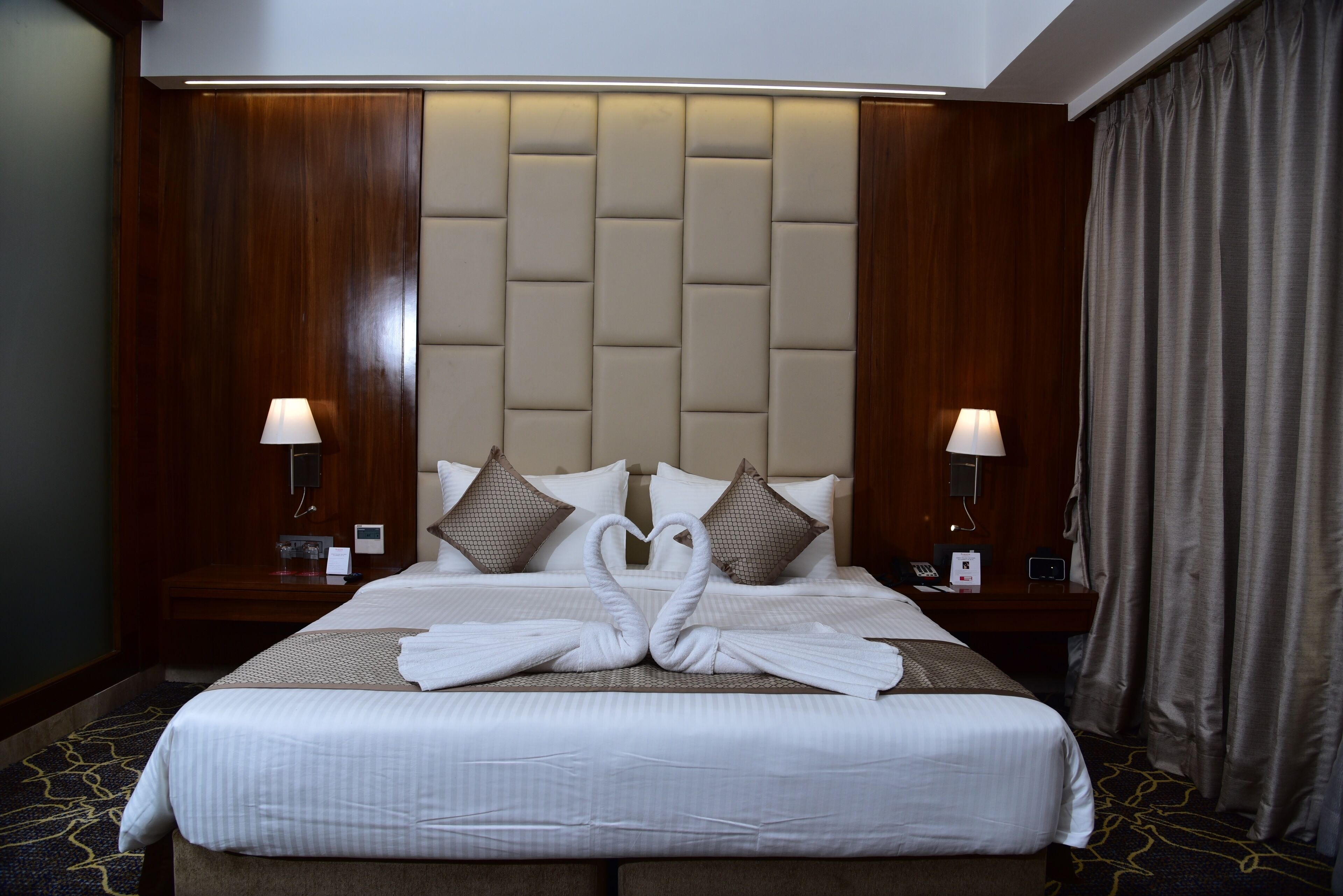 A hotel room with a large bed, two bedside tables with lamps, a headboard, and a curtain.