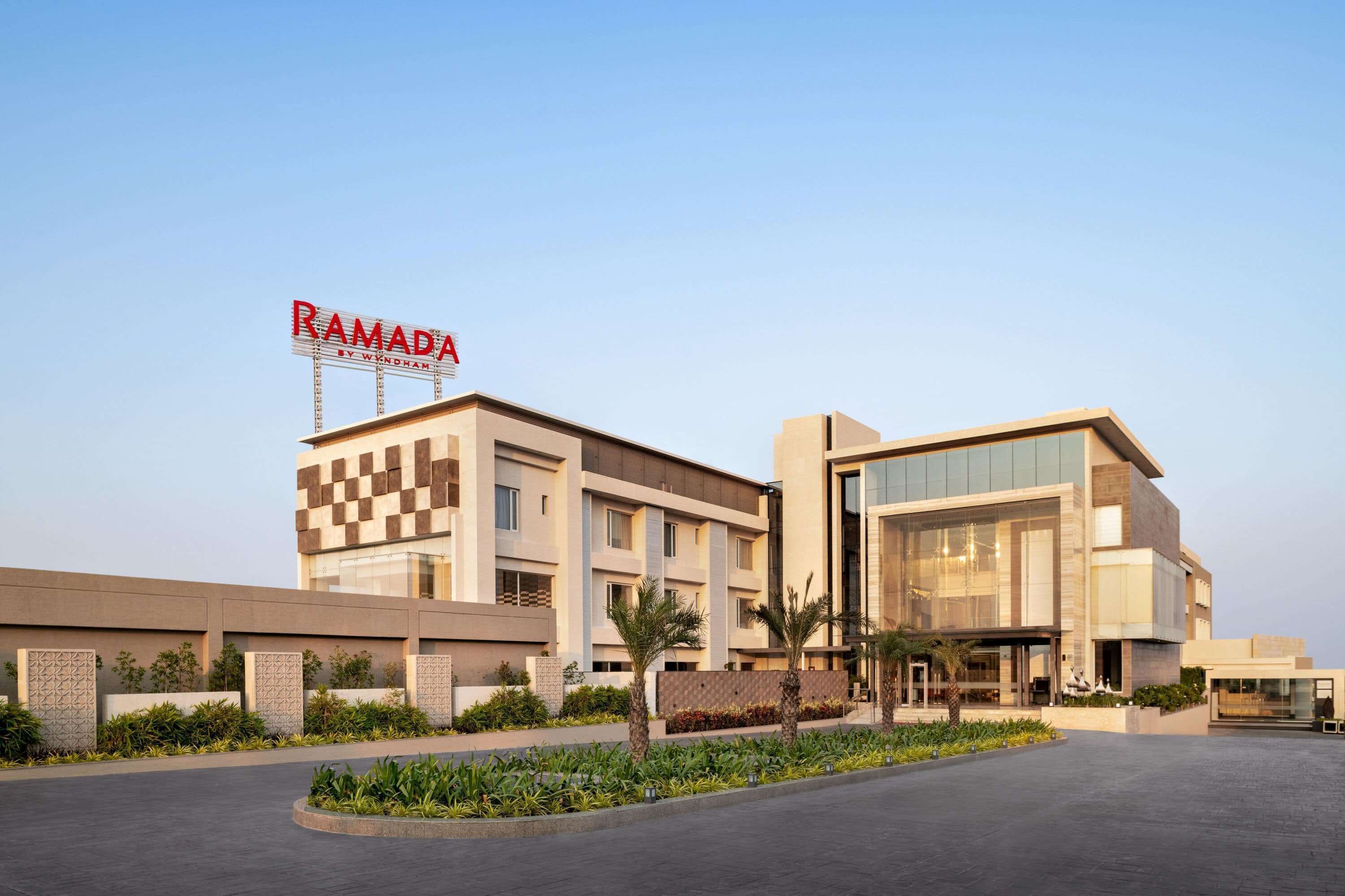 Un hotel moderno con fachada a cuadros, un letrero destacado que dice "RAMADA" y un patio ajardinado.
