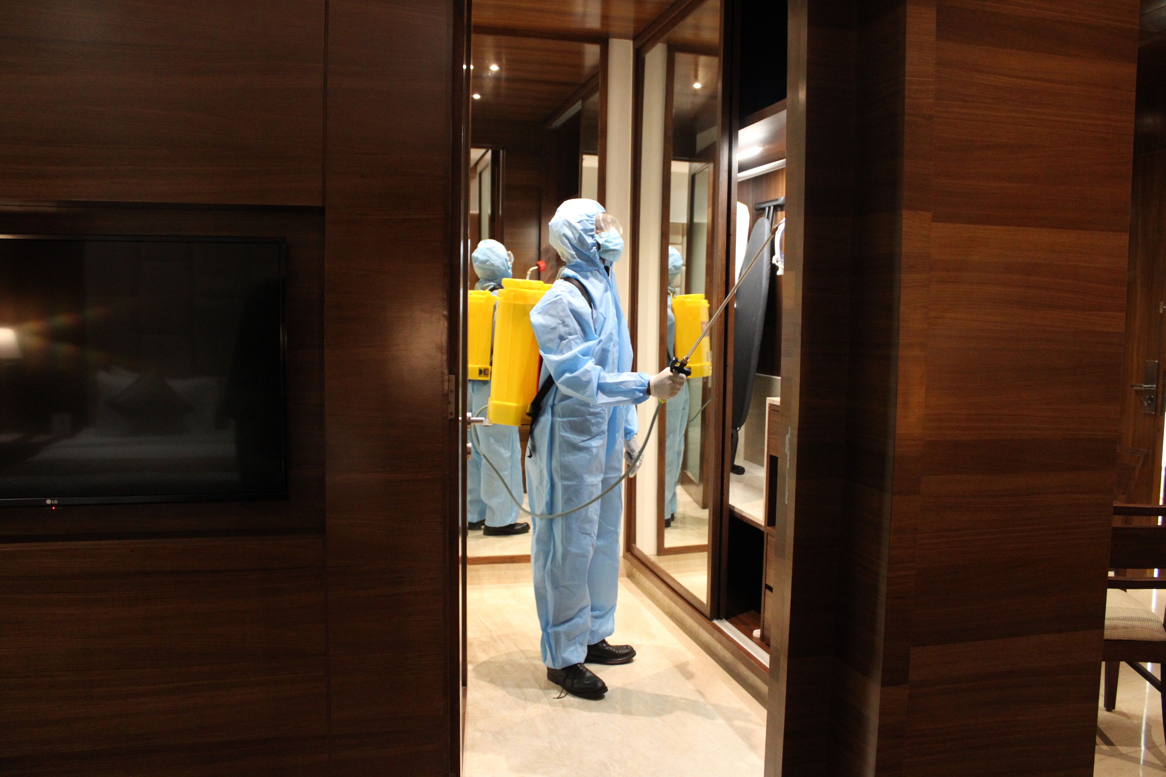 A person in a protective suit spraying disinfectant in a hallway with wooden paneling and a television mounted on the wall.