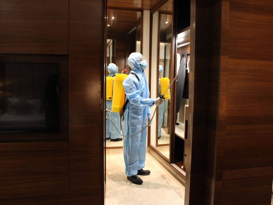 A person in a protective suit spraying disinfectant in a hallway with wooden paneling and a television mounted on the wall.