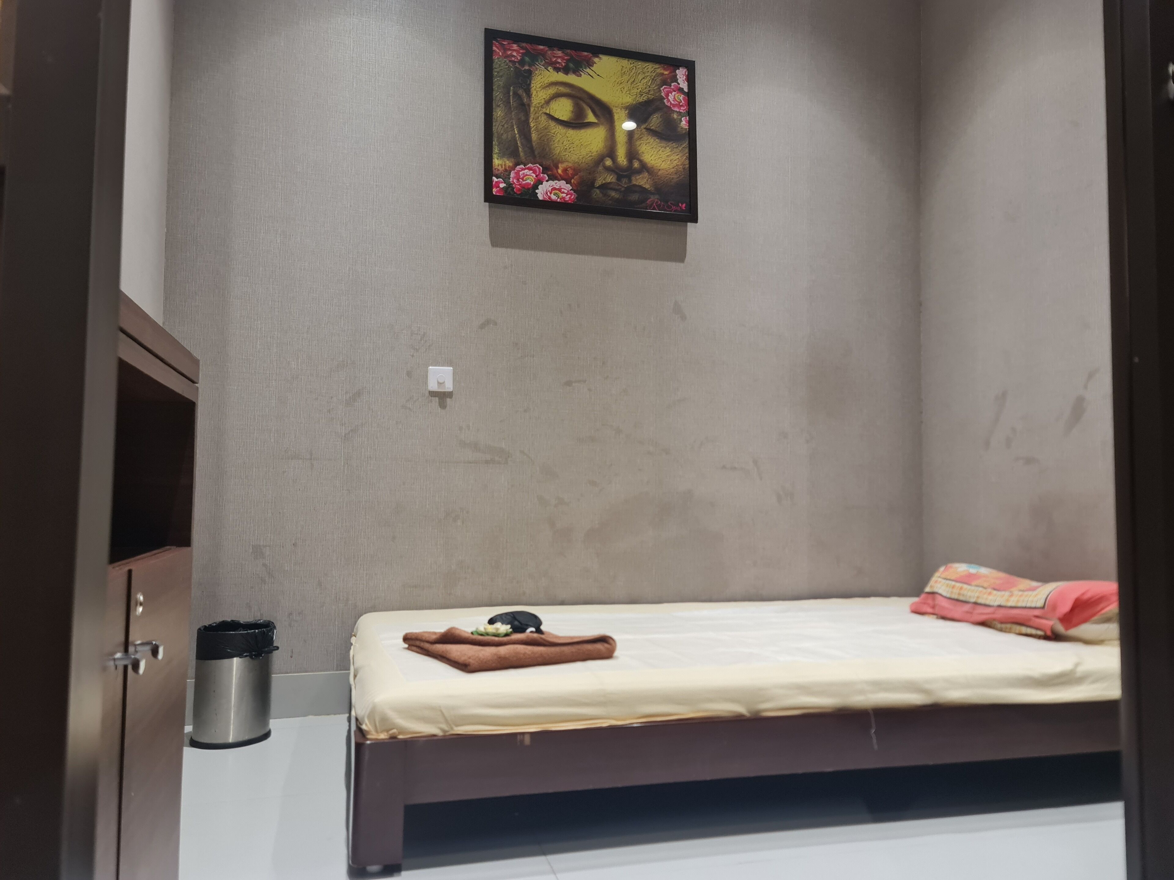 Habitació petita amb un llit individual, una paret sense decoració, un quadre, un cubell de brossa i una porta.
