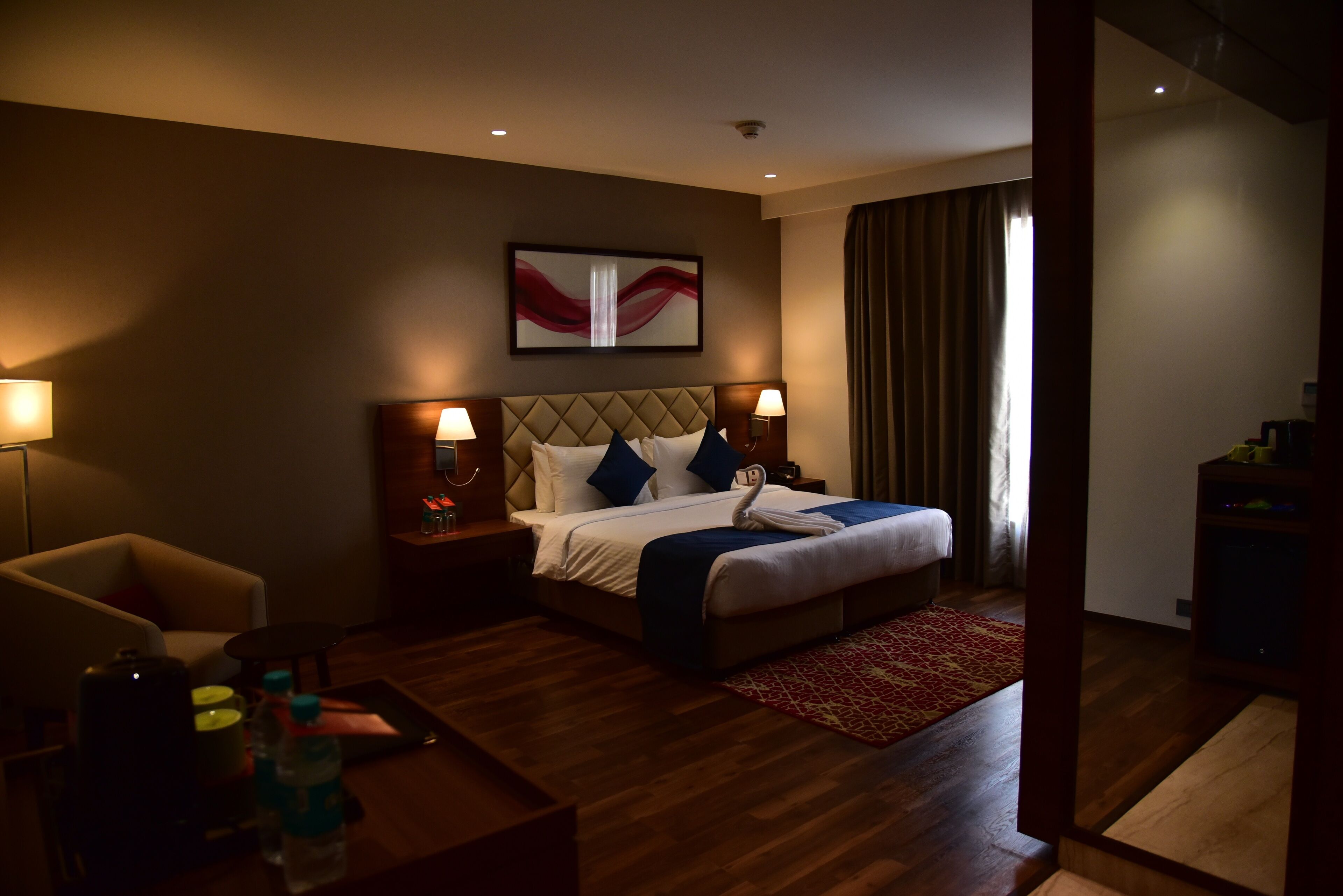 A hotel room with a bed, two bedside tables, a chair, a coffee table, and a red patterned rug.