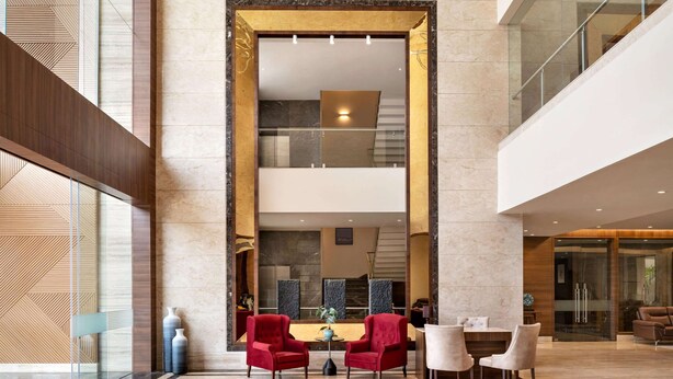 Eine geräumige Lobby mit einer Rezeption, zwei roten Sesseln, einem weißen Tisch und einer Treppe im Hintergrund.