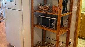 Cocina compacta con refrigerador blanco, microondas y estante de madera con cestas y utensilios de cocina.