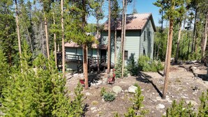 A cabin surrounded by tall pine trees, with a deck and a small outdoor seating area.