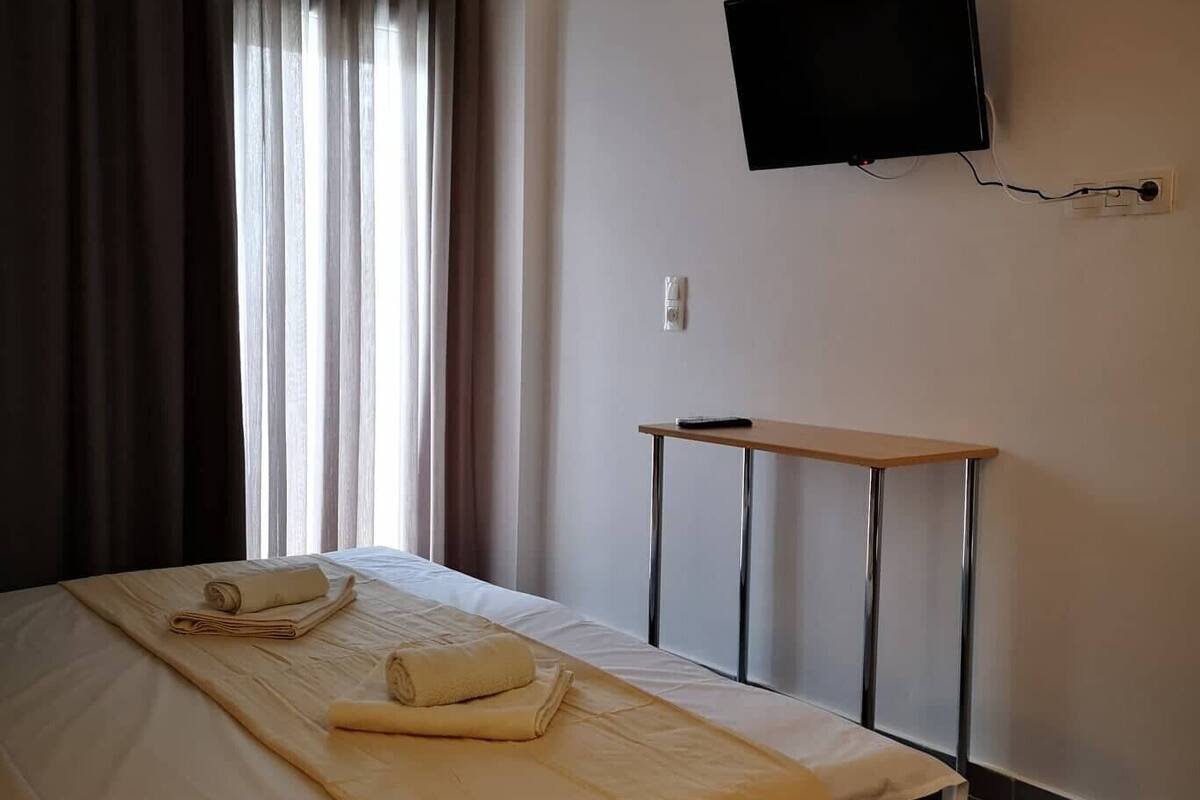 Hotellihuone, jossa on sänky, seinälle asennettu televisio, pieni pöytä ja ikkuna, jossa on verhot.