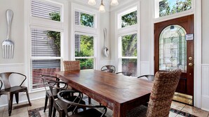 A dining area with a wooden table and chairs, large windows with blinds, and a decorative door with a glass panel. - Heated Pool Access | 5*Clean | Flex Cancelation (Savannah)