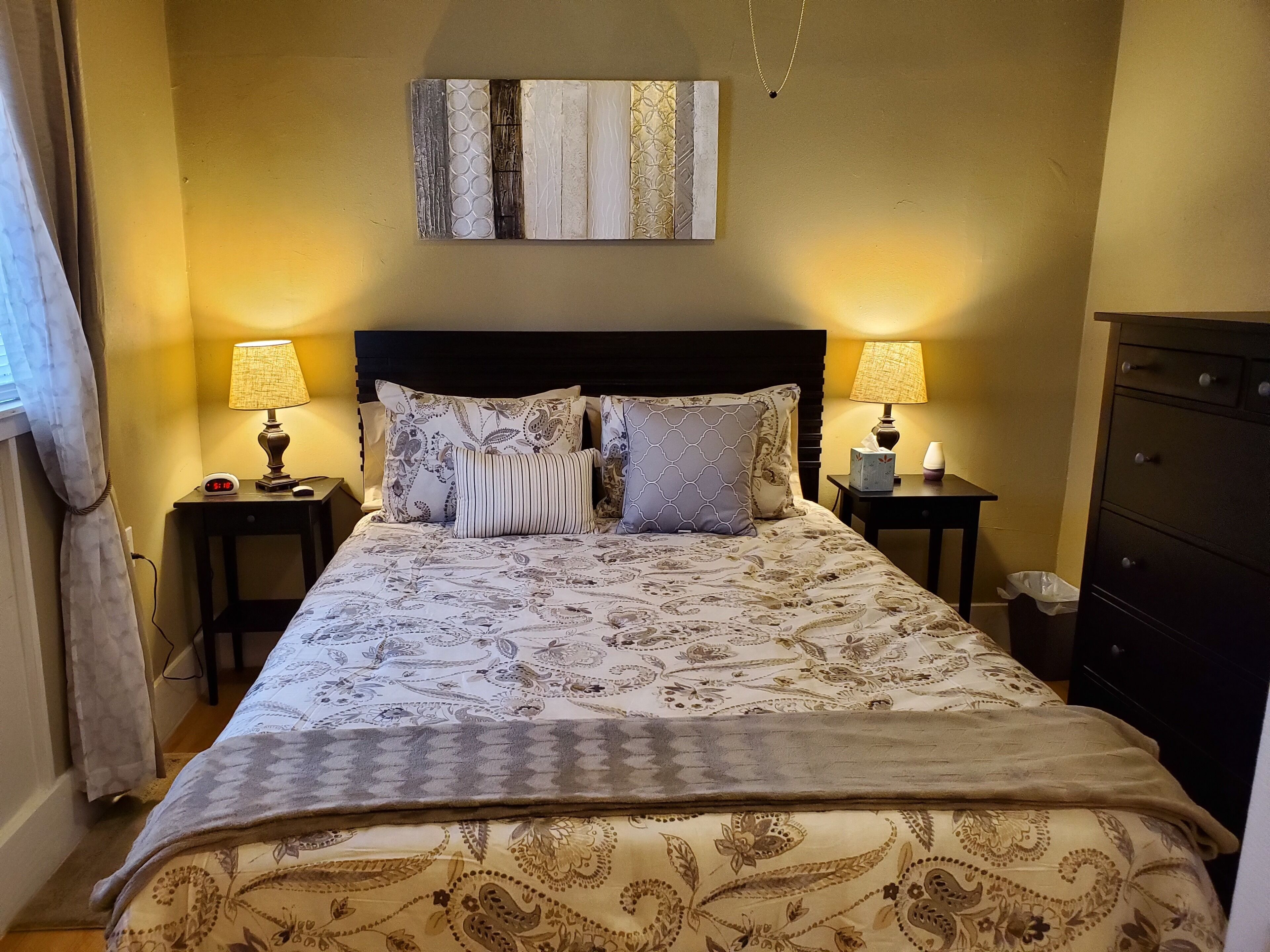 A neatly made bed with patterned bedding, two bedside lamps, a clock, and a wall-mounted artwork.
