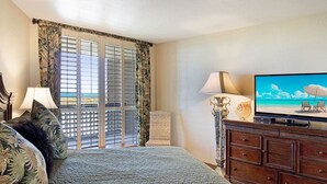 Habitación de hotel con cama, televisor que muestra una escena de playa, cómoda de madera y vista al océano a través de una ventana con cortinas tropicales.