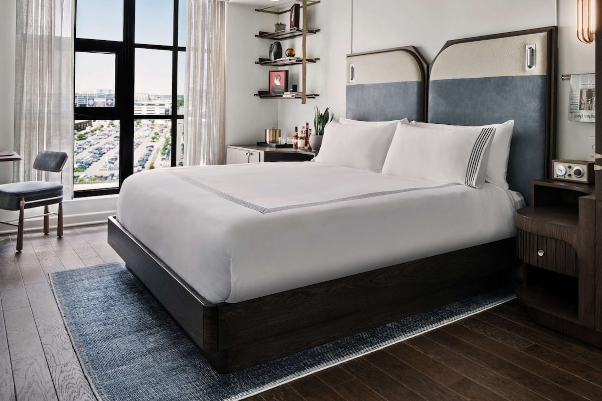 A modern hotel room with a large bed, a desk, and a view of the city through large windows.