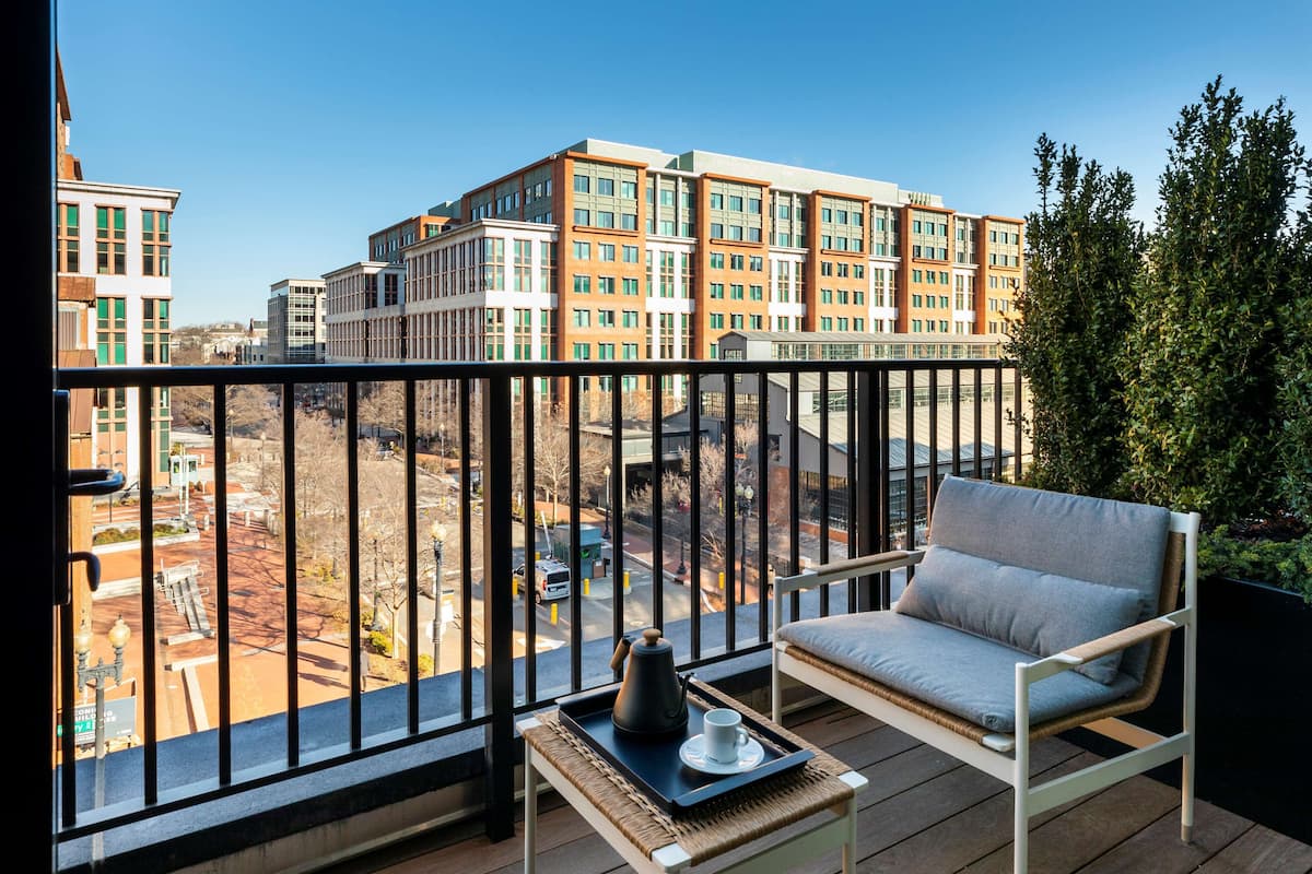 A balcony with a wicker chair and a small table, overlooking a construction site and modern buildings.