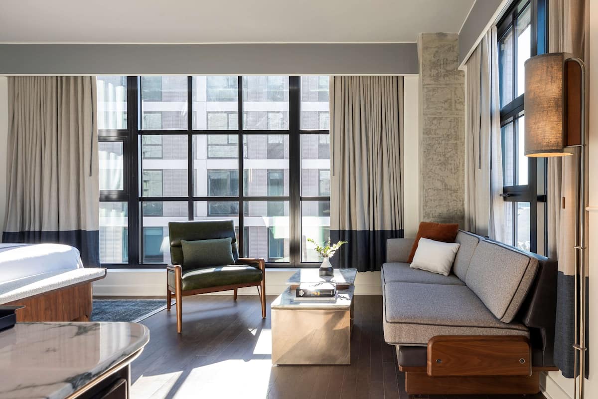 A modern living room with a sofa, armchair, and coffee table, large windows with curtains, and a view of buildings outside.