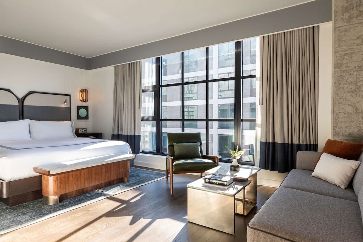 A modern hotel room with a large bed, a green armchair, a grey sofa, and a coffee table.