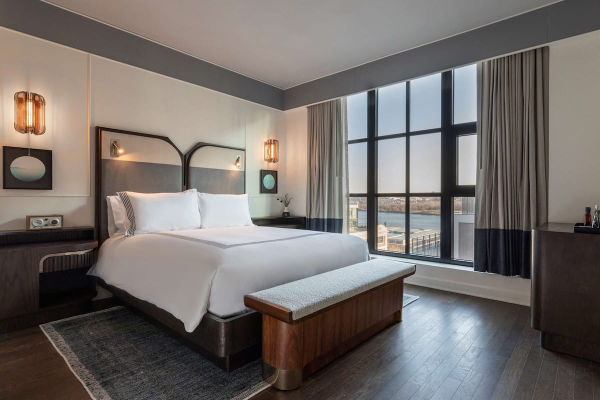 A modern hotel room with a large bed, a bench, a nightstand, and a view of the city through large windows.