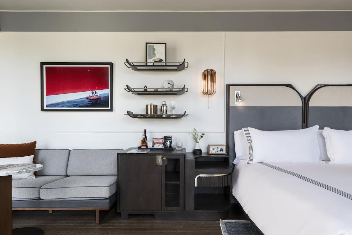 A modern hotel room with a bed, a gray sofa, a small desk with a lamp, and wall-mounted shelves.