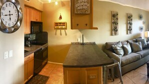 A living room with a sofa, coffee table, and fireplace. There is a kitchen area with a stove and cabinets. The room has a clock on the wall and a bookshelf with books. - Beautiful Updated Condo With Direct River View From Balcony!  We Love Dogs! (New Braunfels)