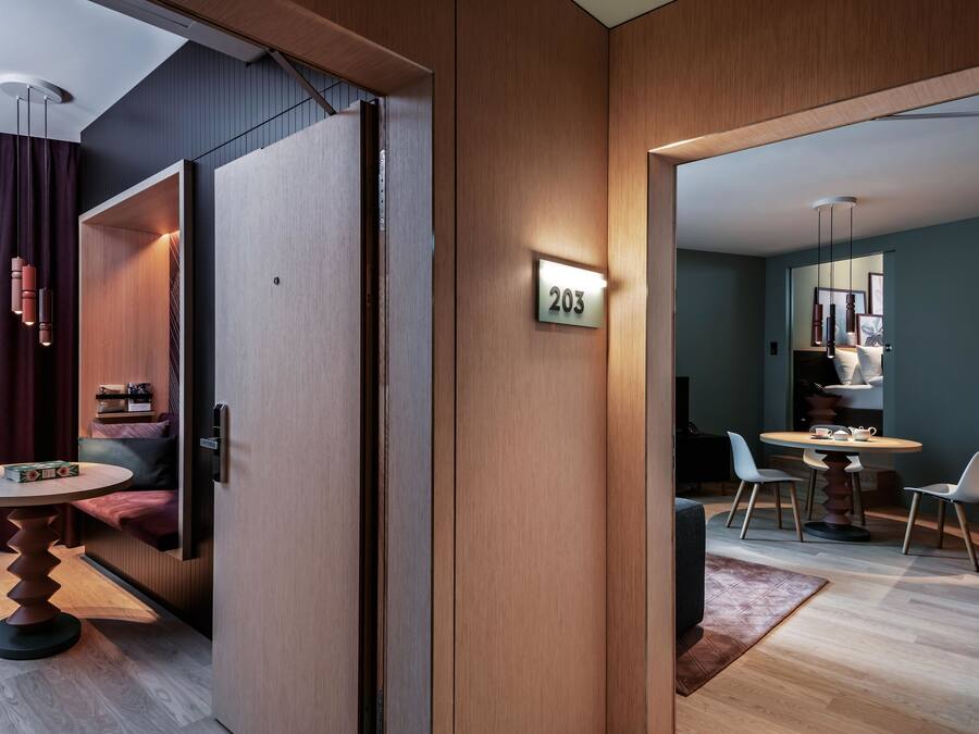 Une chambre d’hôtel moderne avec un intérieur en bois, un coin repas comprenant une table ronde et des chaises, et un numéro de chambre bien visible.