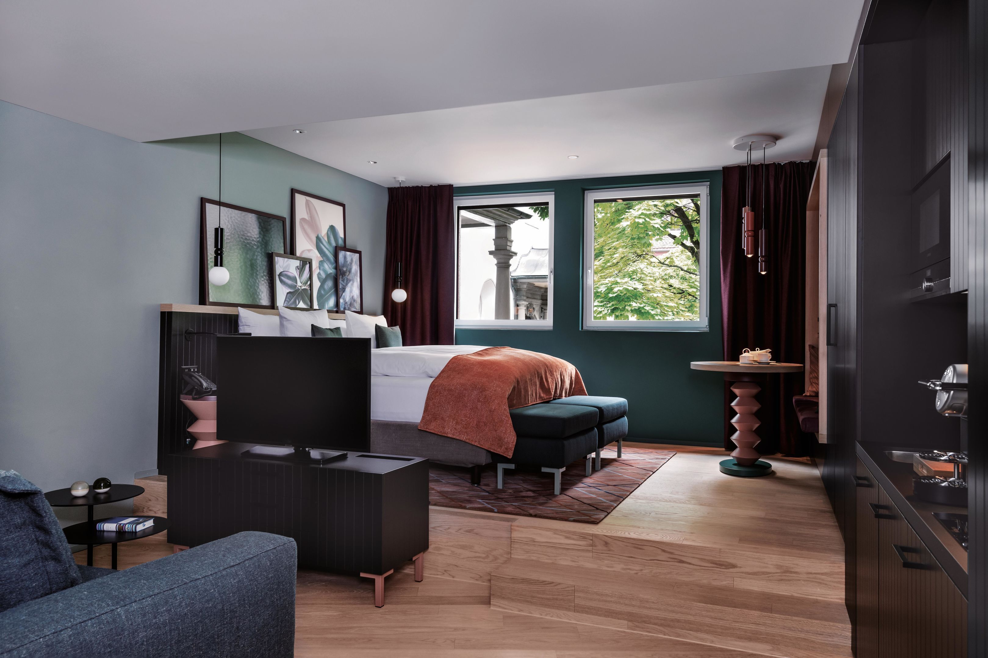 Ein modernes Schlafzimmer mit Bett, Schreibtisch, Stuhl, Sofa und Fenster mit Blick auf Grünflächen.