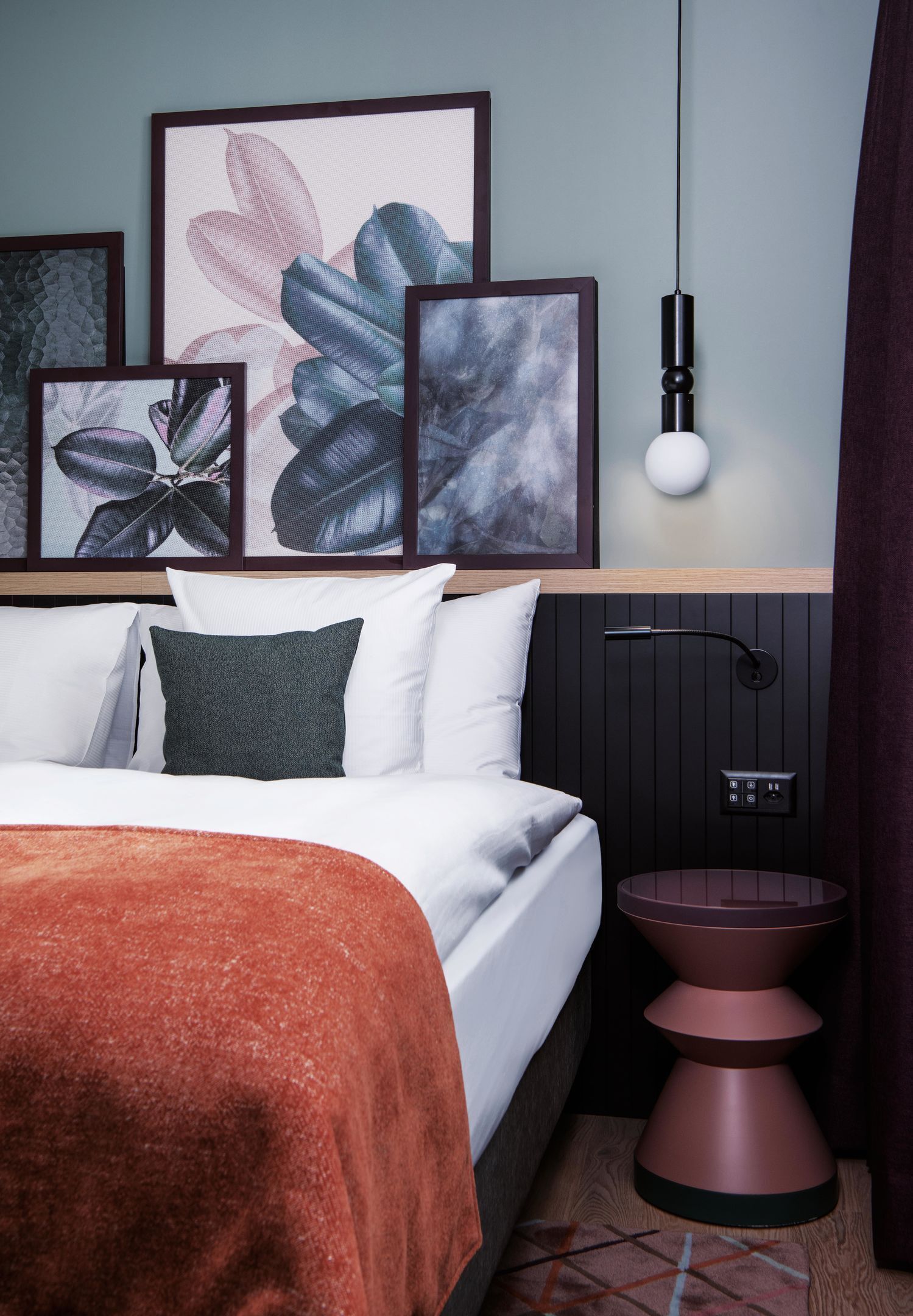 A bedroom with a bed, pillows, a bedside table, a wall-mounted light, and framed artwork.