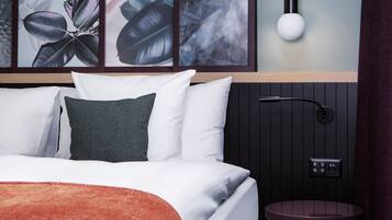 A bedroom with a bed, pillows, a bedside table, a wall-mounted light, and framed artwork.