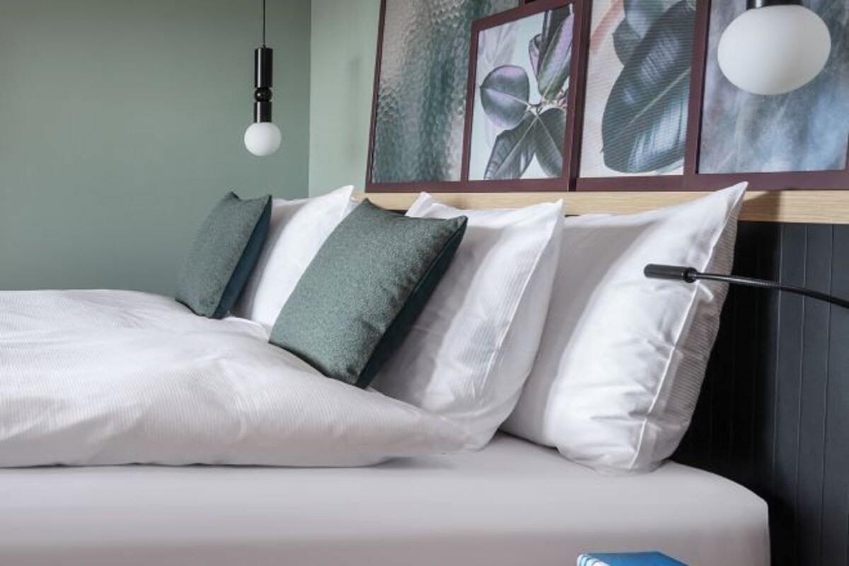Una cama bien hecha con almohadas blancas y verdes, una pared blanca y una obra de arte botánica enmarcada encima.