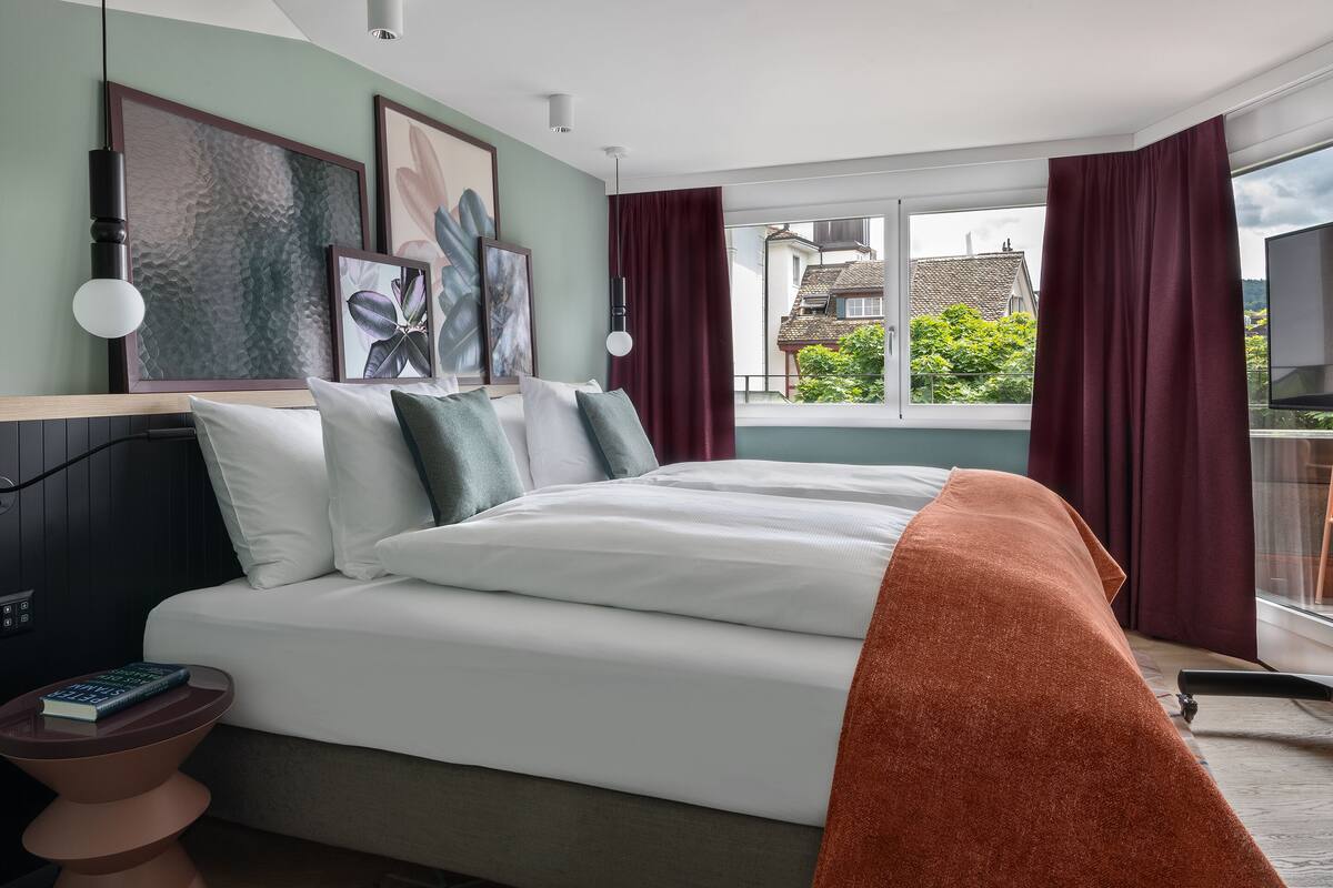 Ein modernes Schlafzimmer mit einem großen Bett, einem Nachttisch mit einem Buch und Blick auf Grünflächen und Gebäude.