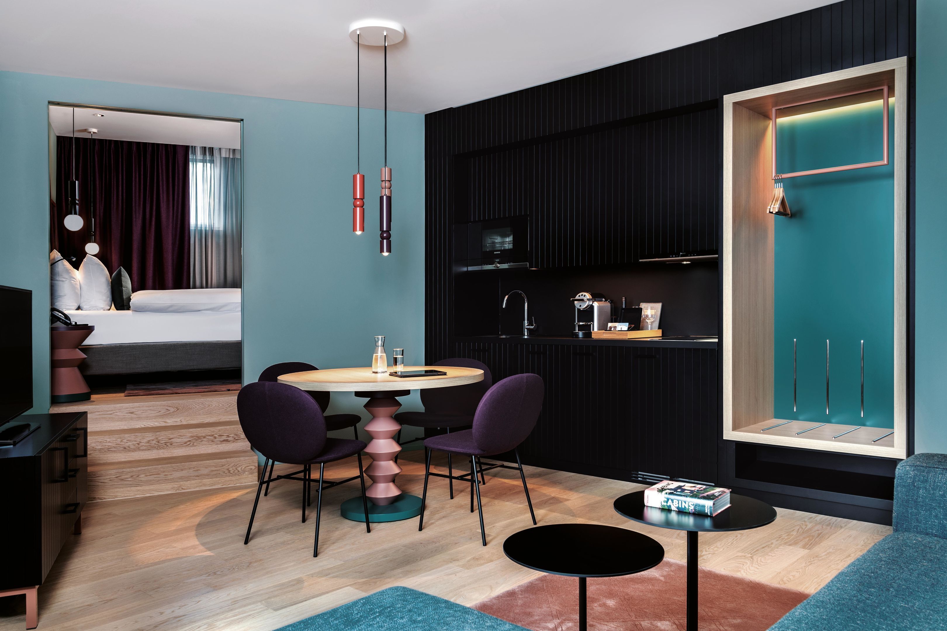 A modern hotel room with a dining area, a bed, a TV, and a kitchen area.