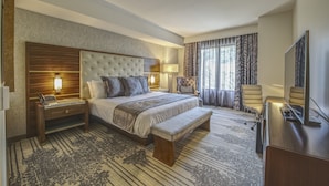 A hotel room with a large bed, a wooden nightstand, a bench, a desk with a TV, and a chair. - Legacy Resort Hotel & Spa (San Diego)