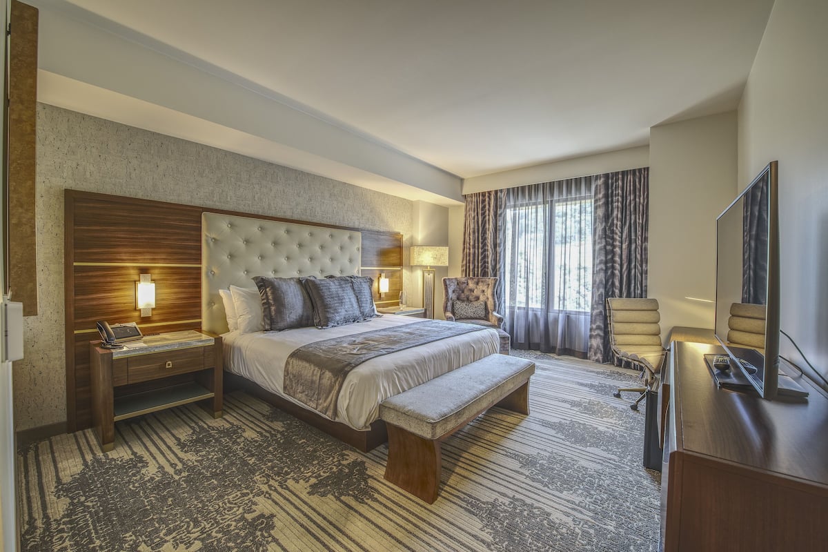 A hotel room with a large bed, a wooden nightstand, a bench, a desk with a TV, and a chair.