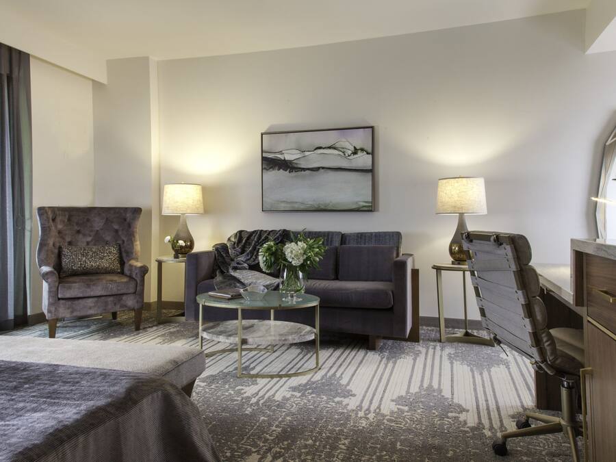 A hotel room with a bed, a grey armchair, a sofa, a coffee table, and a desk with a lamp. There is a painting on the wall and a window with curtains.