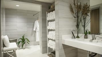 A modern bathroom with a large mirror, white sink, and towel storage. There is a shower area with a glass door and a white bath chair.