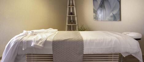 Una cama bien tendida con una manta estampada, un taburete pequeño, un estante en la pared con plantas y un cuadro.