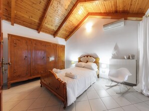 A bedroom with a wooden ceiling, a large wardrobe, a bed with white linens, a rocking chair, and a radiator. - Villa Eleni-2nd Floor Maisonette with Ocean panorama terrace (Alikanas)