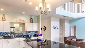 A modern kitchen with white cabinets, a black countertop, and a chandelier. There is a dining area with a brown leather couch and a teal bench. - 8BR lakefront Disney home w themed rms HarryPotter Frozen ToyStory Mario Royal (Kissimmee)