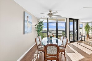 Un coin repas avec une table en verre, des chaises en osier, un ventilateur de plafond et une vue sur l’océan.